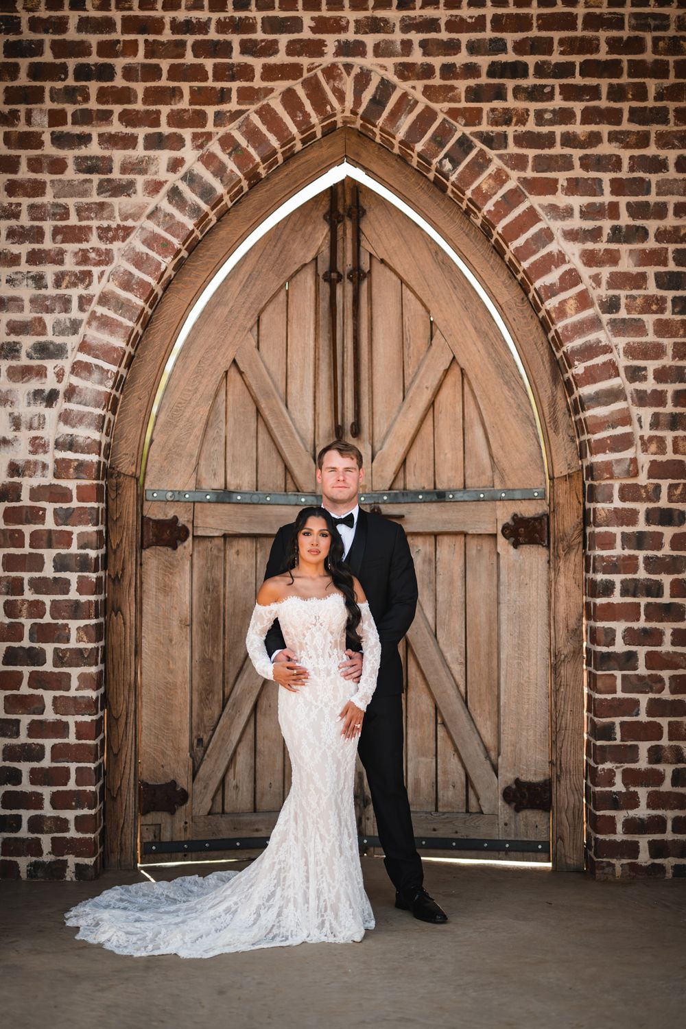 Bride in lace gown and groom in tuxedo pose before Gothic arched wooden doors in brick wall.