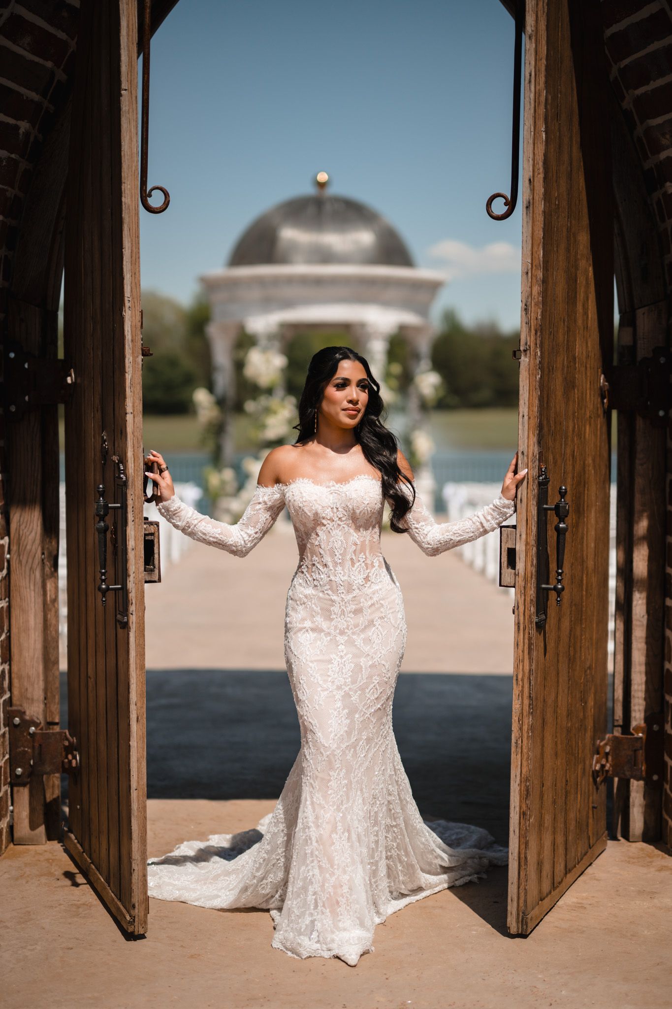 Bride in fitted off-shoulder lace wedding gown stands in open wooden doors with a gazebo and lake behind.