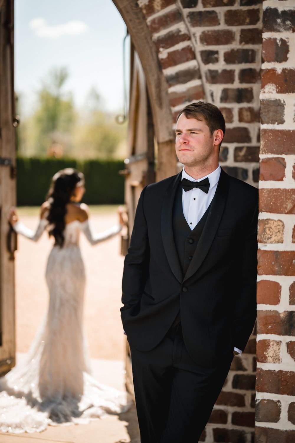 Groom in black tuxedo leans against brick wall while bride in lace gown stands in doorway behind him.