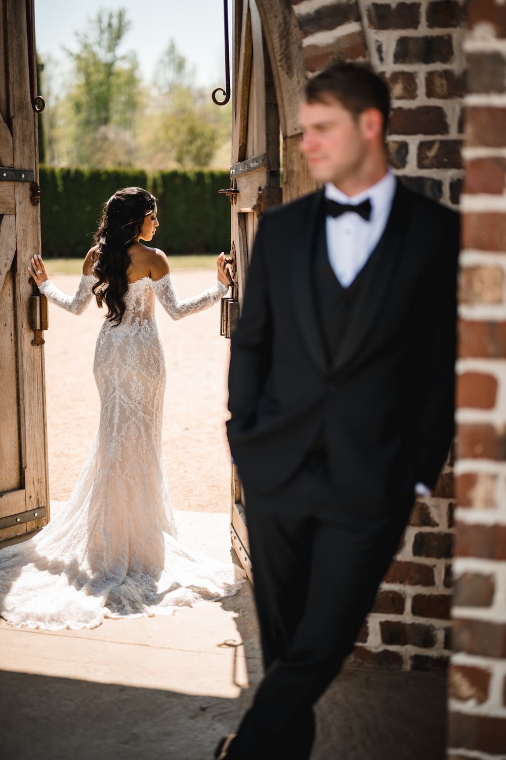Bride in lace gown opens barn doors as groom in tuxedo stands nearby in sunlit outdoor wedding.