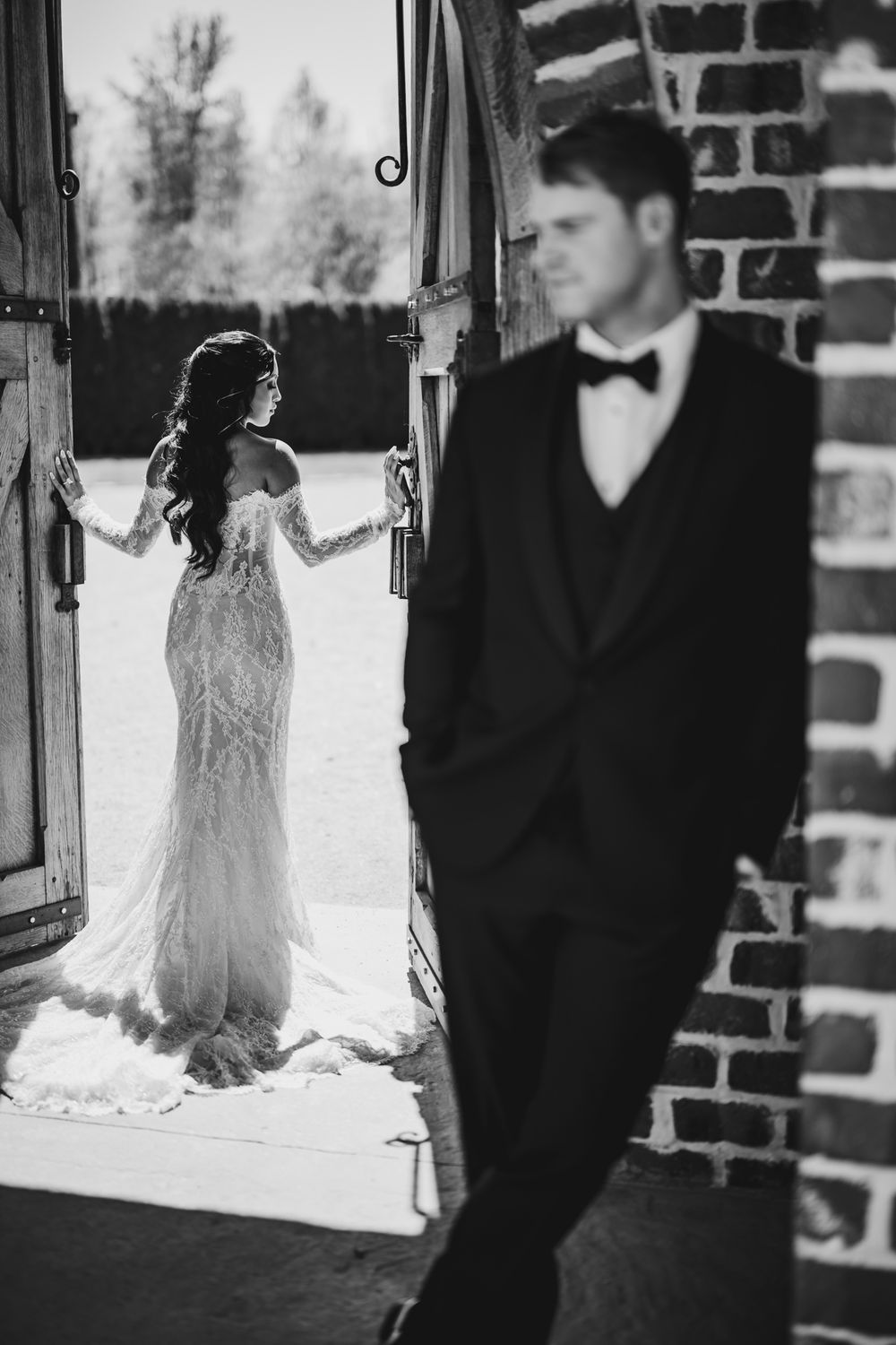 Black and white photo of bride in lace gown at doorway with groom in tuxedo leaning on brick wall.