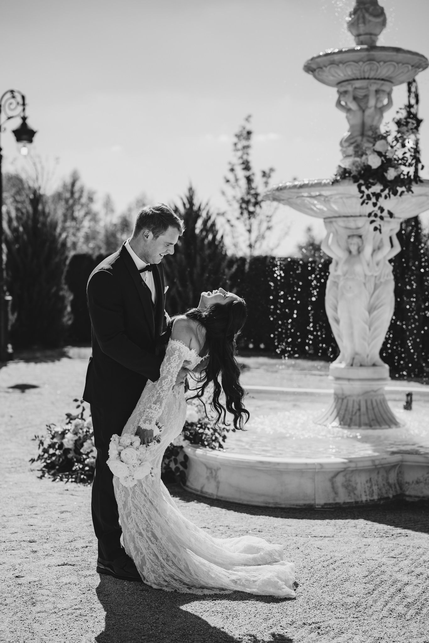 Black and white photo of bride and groom sharing romantic dip near ornate stone fountain.