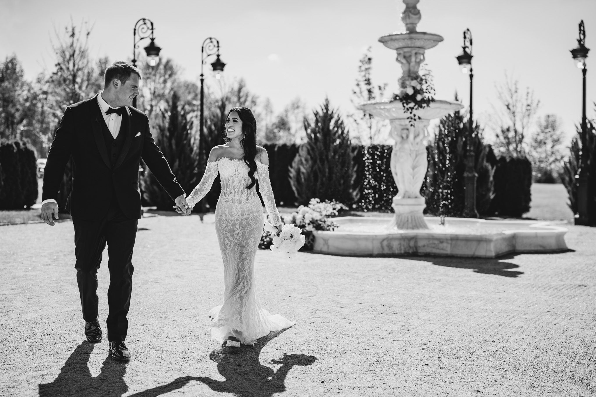 Black and white photo of bride and groom walking near a fountain at their venue.