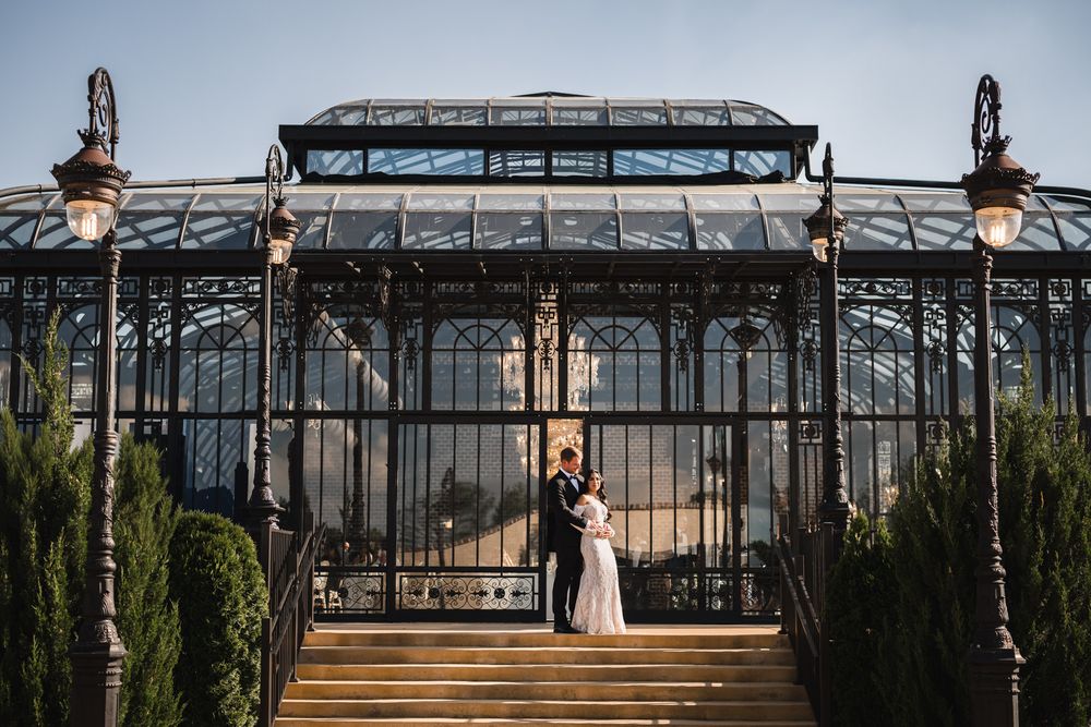 Couple embraces on steps before an elegant Victorian glass conservatory at golden hour.