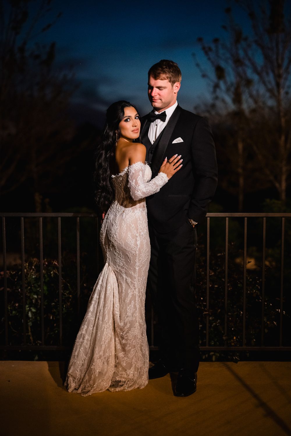 Elegant bride and groom in formal wedding attire pose together on terrace under dramatic twilight sky.