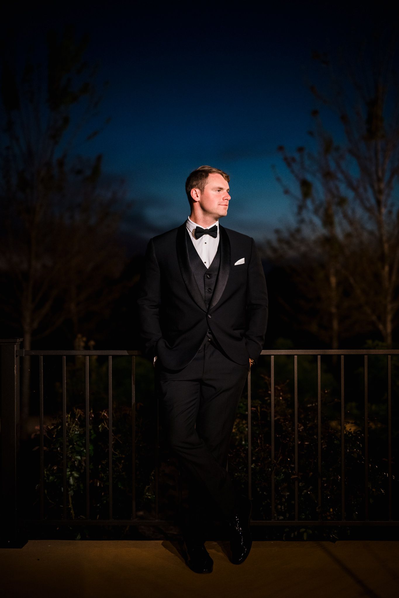 Dapper groom in black tuxedo with bow tie stands confidently against a railing at dusk outdoors.