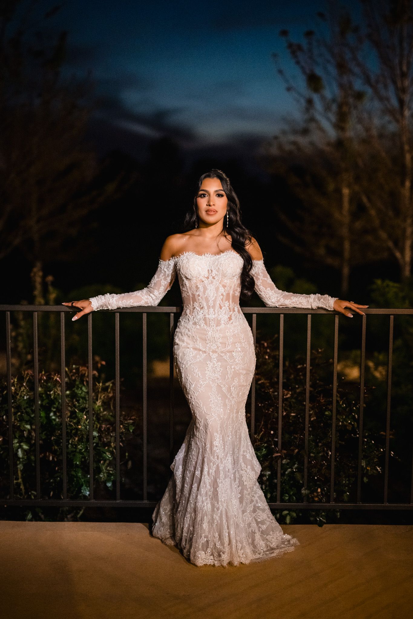 Bride poses gracefully in a fitted lace off-shoulder gown against a railing at dusk.