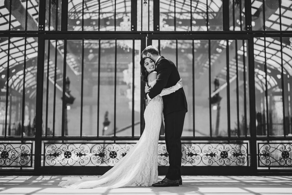Bride and groom embrace in elegant black and white wedding photo inside a grand glass conservatory.