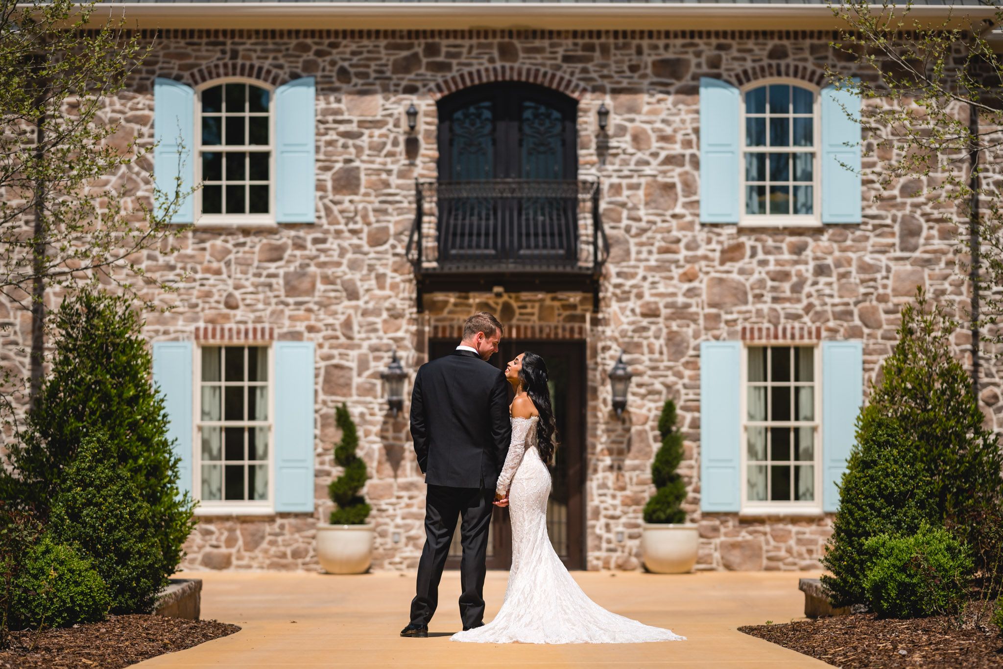 Bride and groom share a romantic moment in front of a stone mansion with blue shutters on their wedding day.