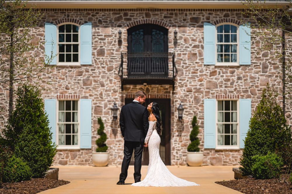 Bride and groom share a romantic moment in front of a stone mansion with blue shutters on their wedding day.