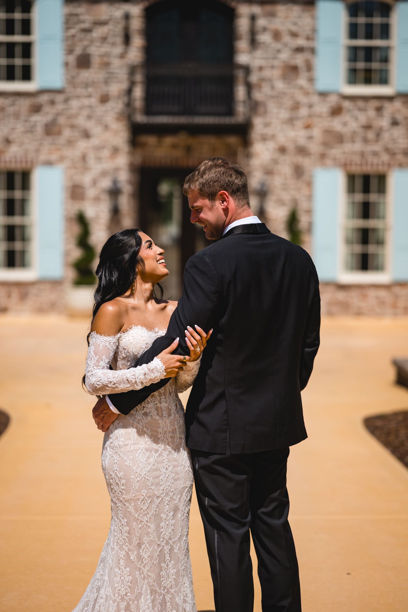 Bride in lace gown embraces groom in black tuxedo outside elegant brick mansion on wedding day.