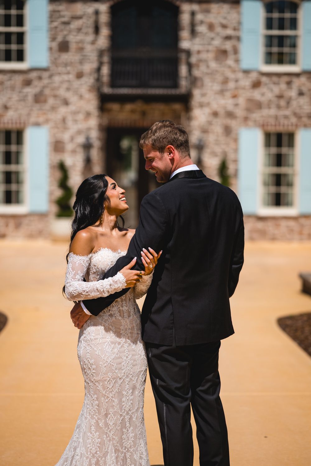 Bride in lace gown embraces groom in black tuxedo outside elegant brick mansion on wedding day.