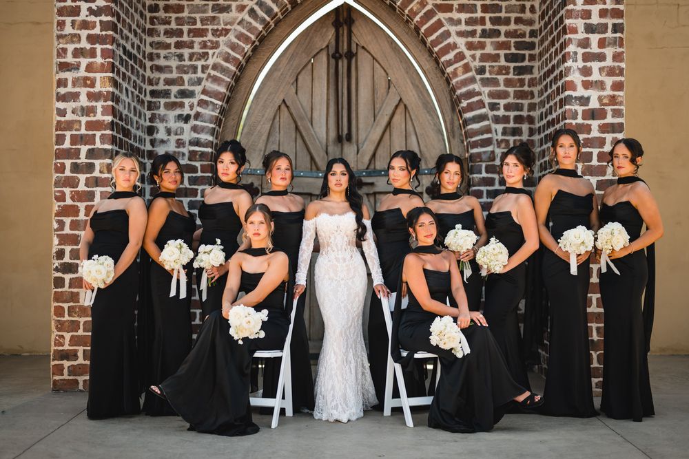 Bride in white gown poses with bridesmaids in black dresses holding white bouquets before a brick church.
