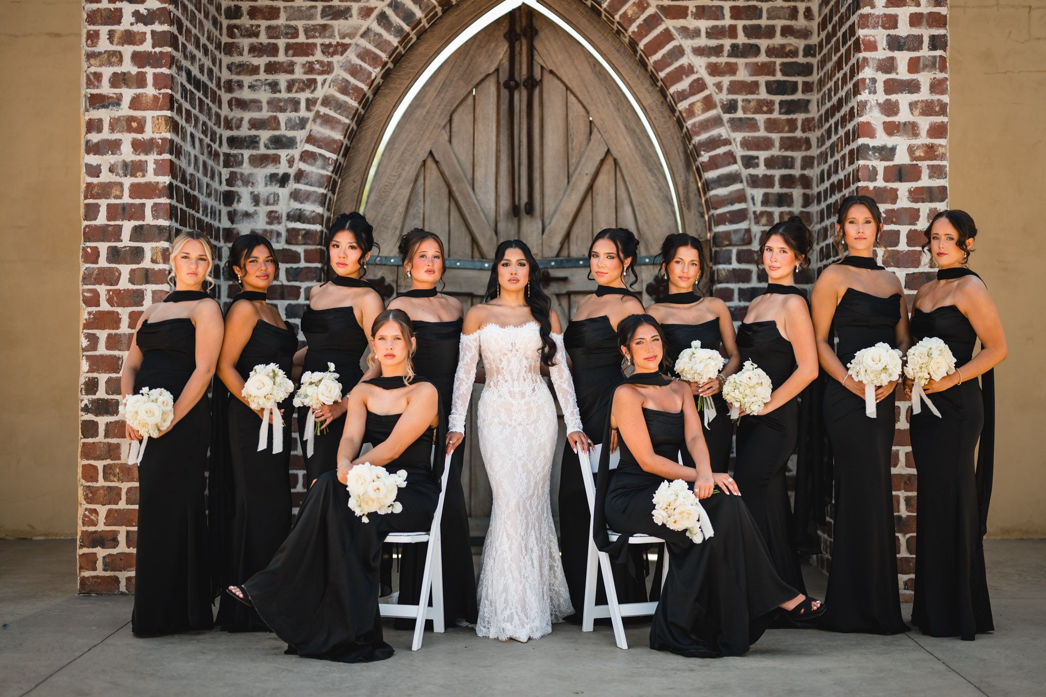 Bride in white gown poses with bridesmaids in black dresses holding white bouquets before a brick church.