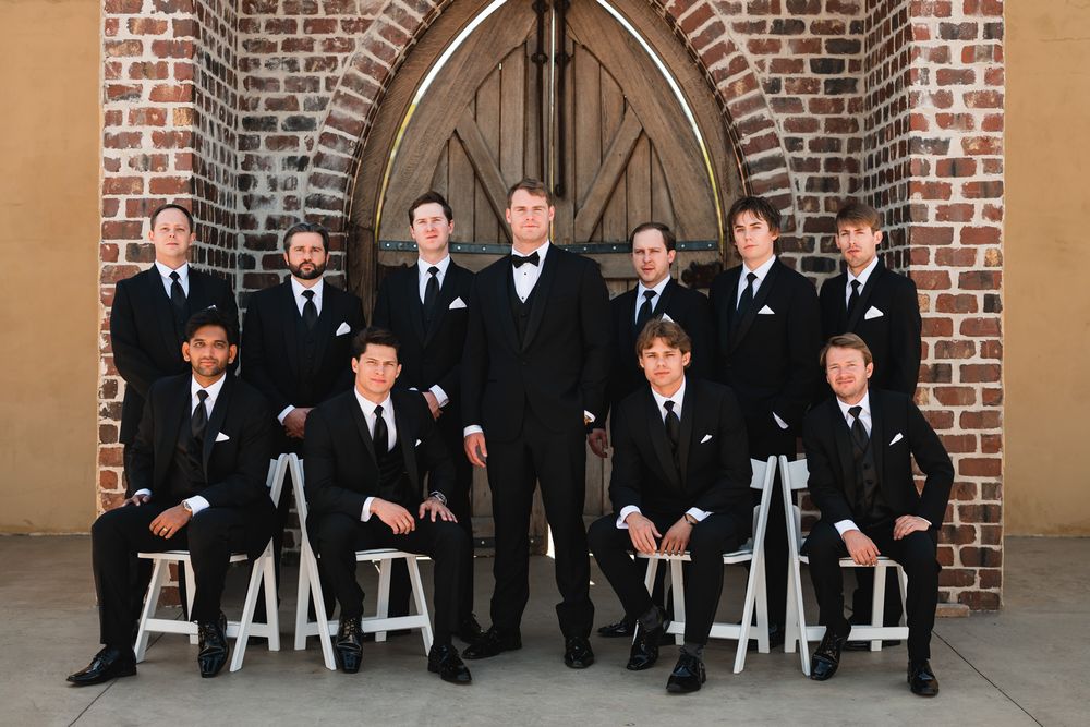 Groom and groomsmen in black tuxedos pose in front of a brick church with arched wooden doors.