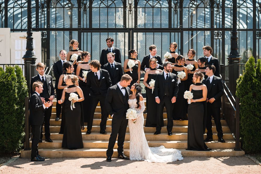 Wedding party in black attire poses on grand steps outside an elegant glass greenhouse venue.