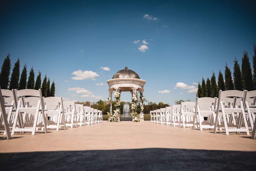 Elegant outdoor wedding ceremony setup with white chairs flanking an aisle leading to a white gazebo altar.