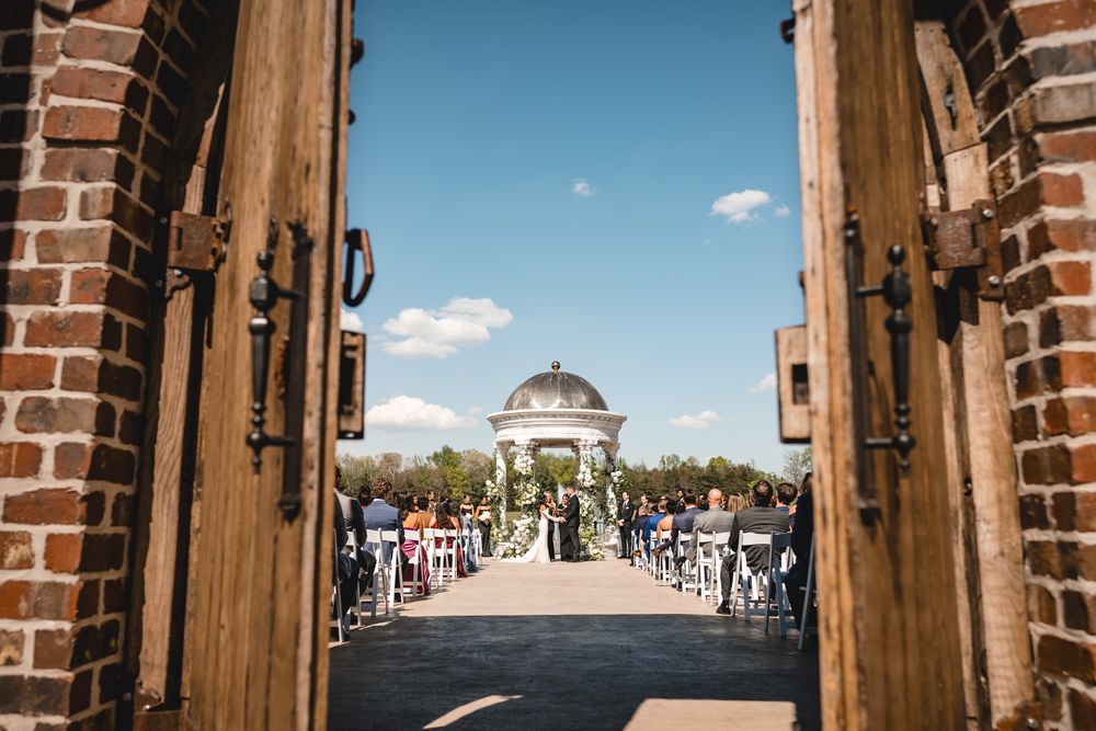 Stunning outdoor wedding ceremony viewed through rustic wooden doors with white gazebo altar.