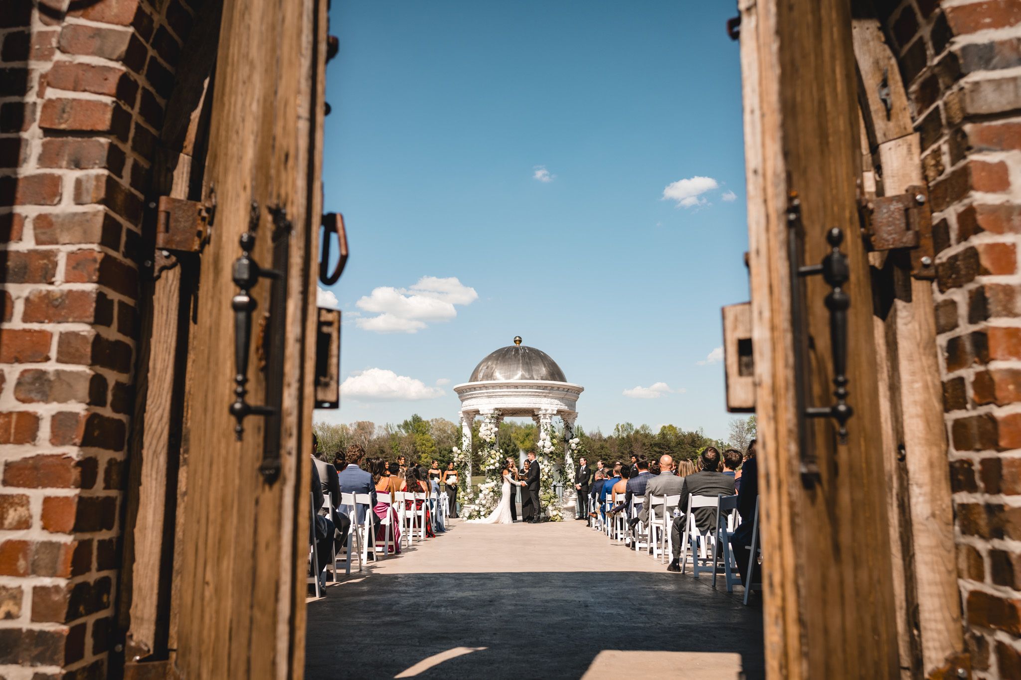 Stunning outdoor wedding ceremony viewed through rustic wooden doors with white gazebo altar.