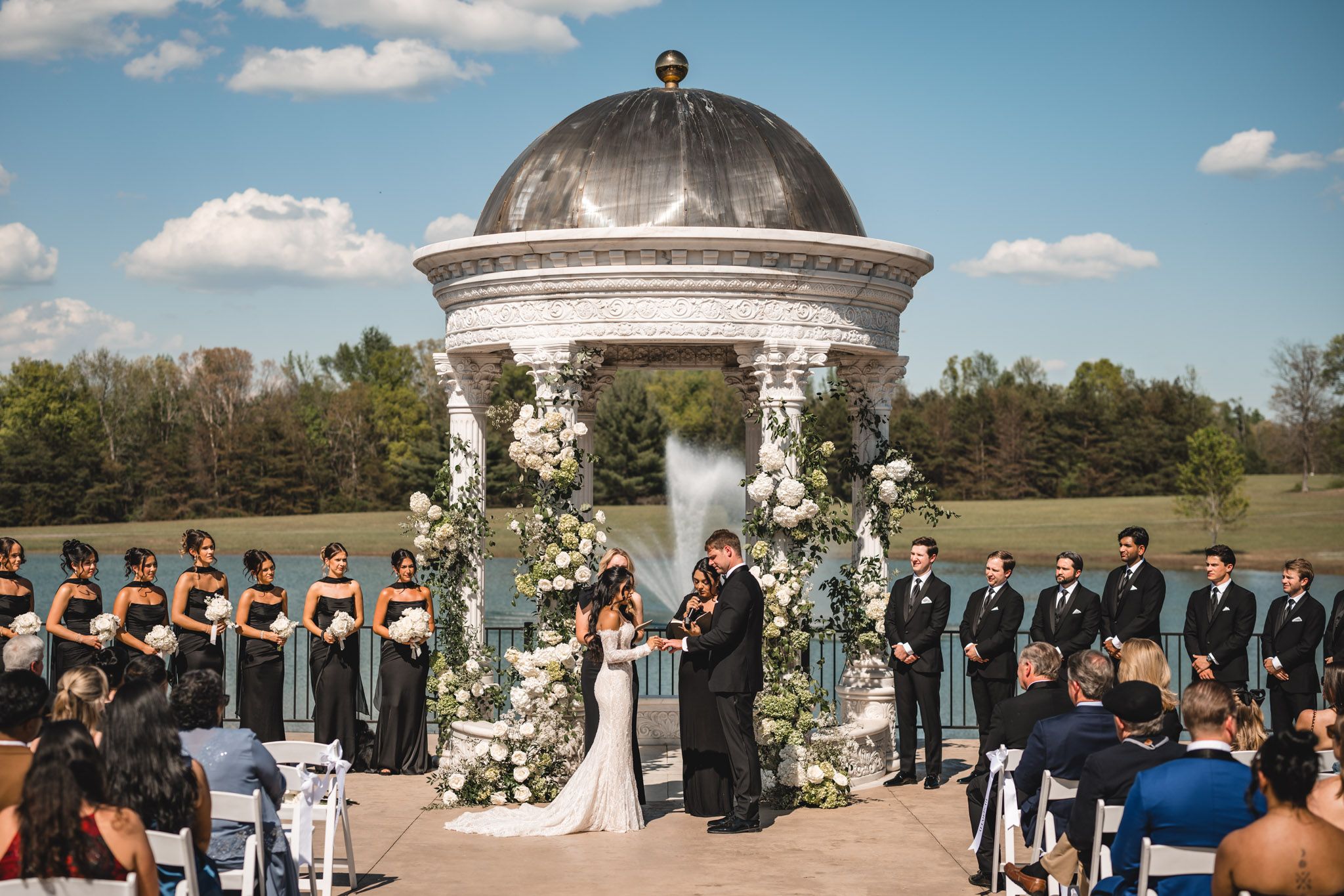 Couple exchanges vows at ornate gazebo with white floral arch, bridal party in black attire.