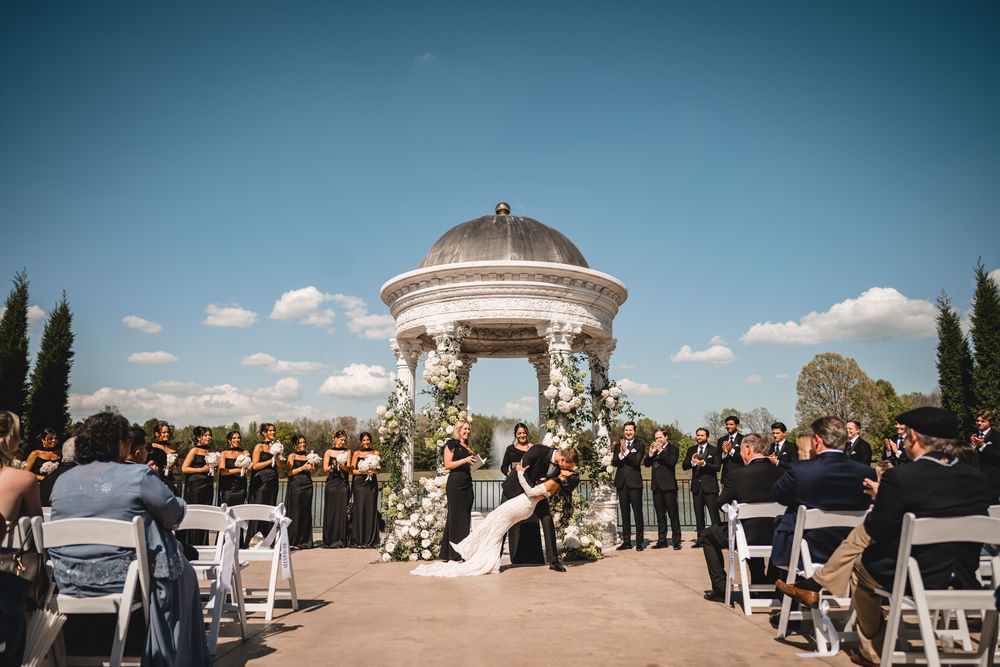 Wedding ceremony at elegant gazebo altar with floral arch, bridesmaids in black, sunny blue sky.
