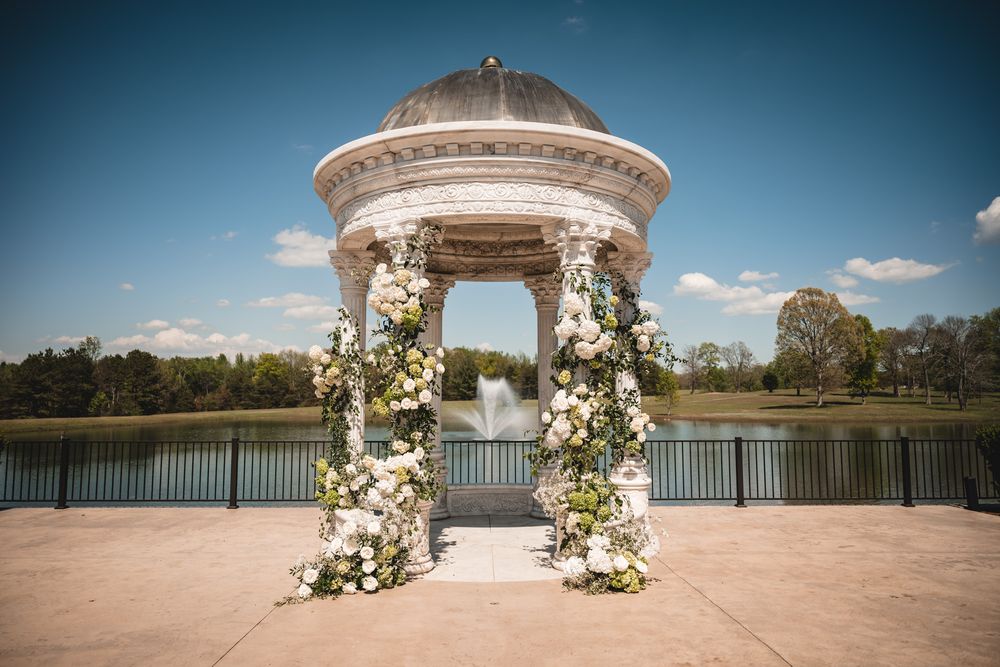 Elegant white gazebo adorned with lush floral arrangements overlooking a serene lake on a sunny day.