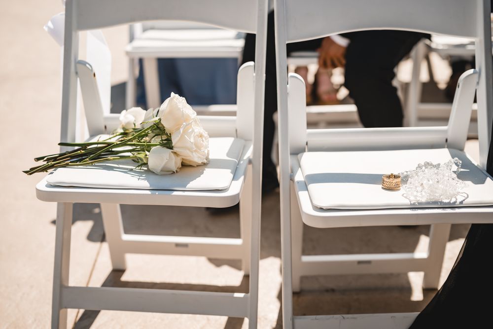 White roses and wedding rings rest on white folding chairs at an outdoor wedding ceremony.