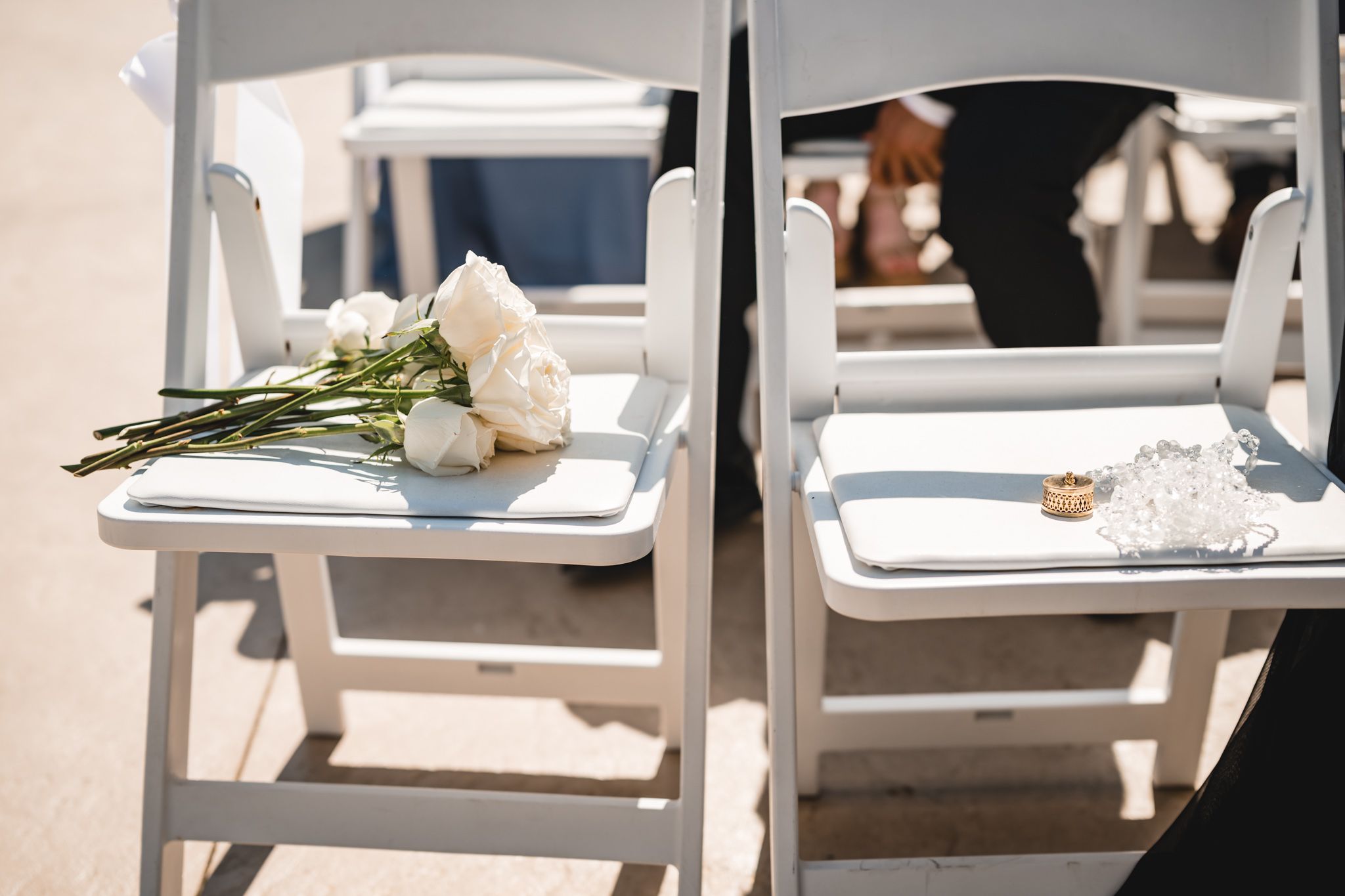 White roses and wedding rings rest on white folding chairs at an outdoor wedding ceremony.