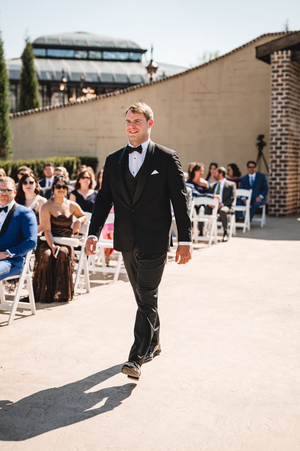 Smiling groom in black tuxedo walks down the aisle at an outdoor wedding ceremony.