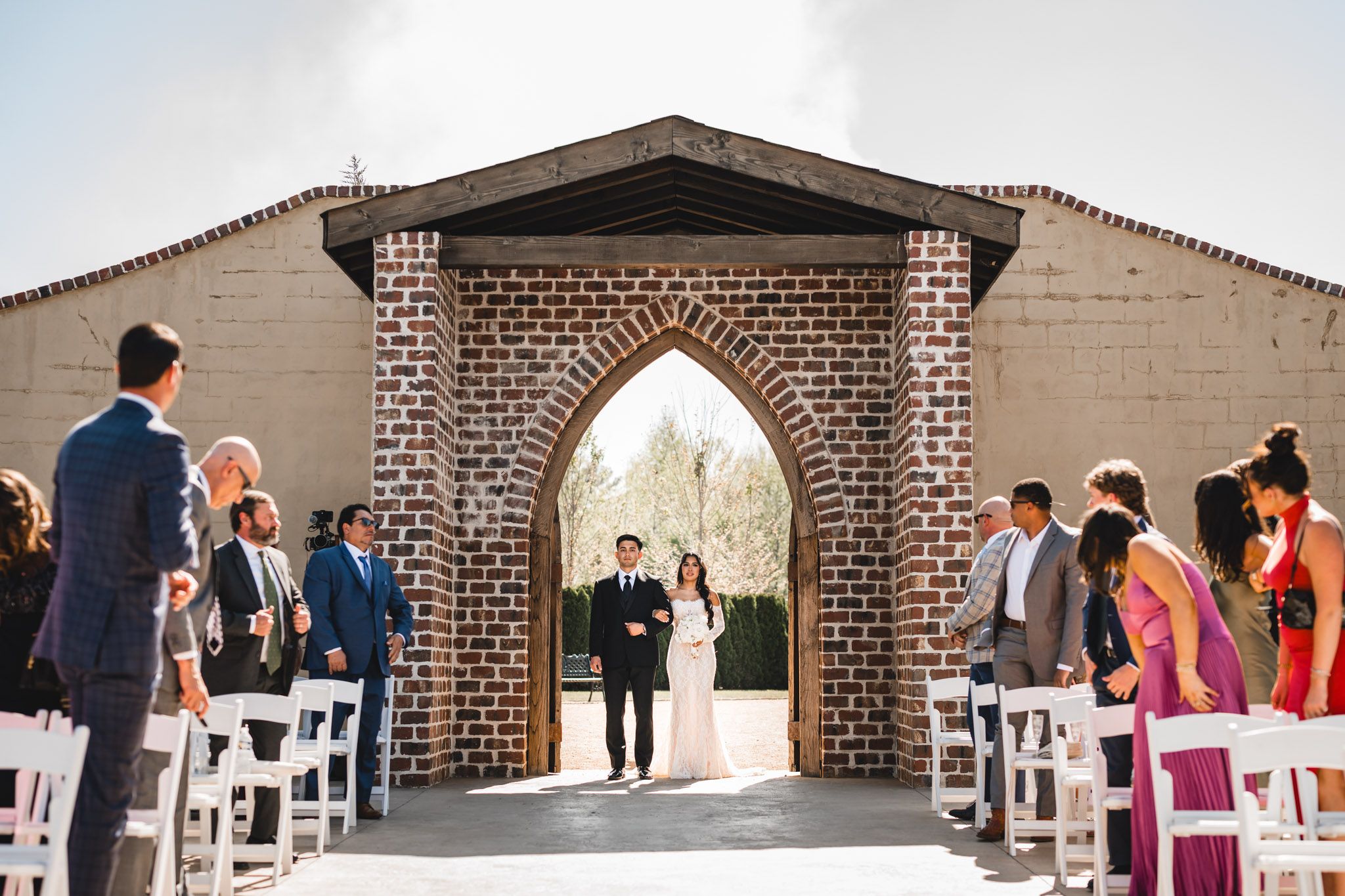 Outdoor wedding ceremony through a Gothic brick arch with guests seated in white chairs.