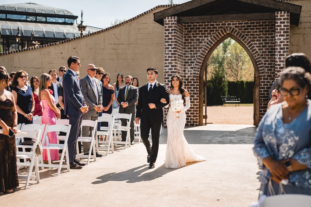 Bride walks down outdoor wedding aisle with escort through elegant stone arch as guests stand.