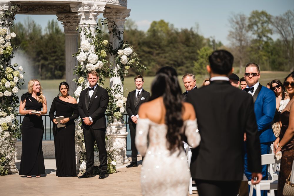 Groom awaits bride at outdoor wedding ceremony beside a floral-decorated gazebo with guests watching.