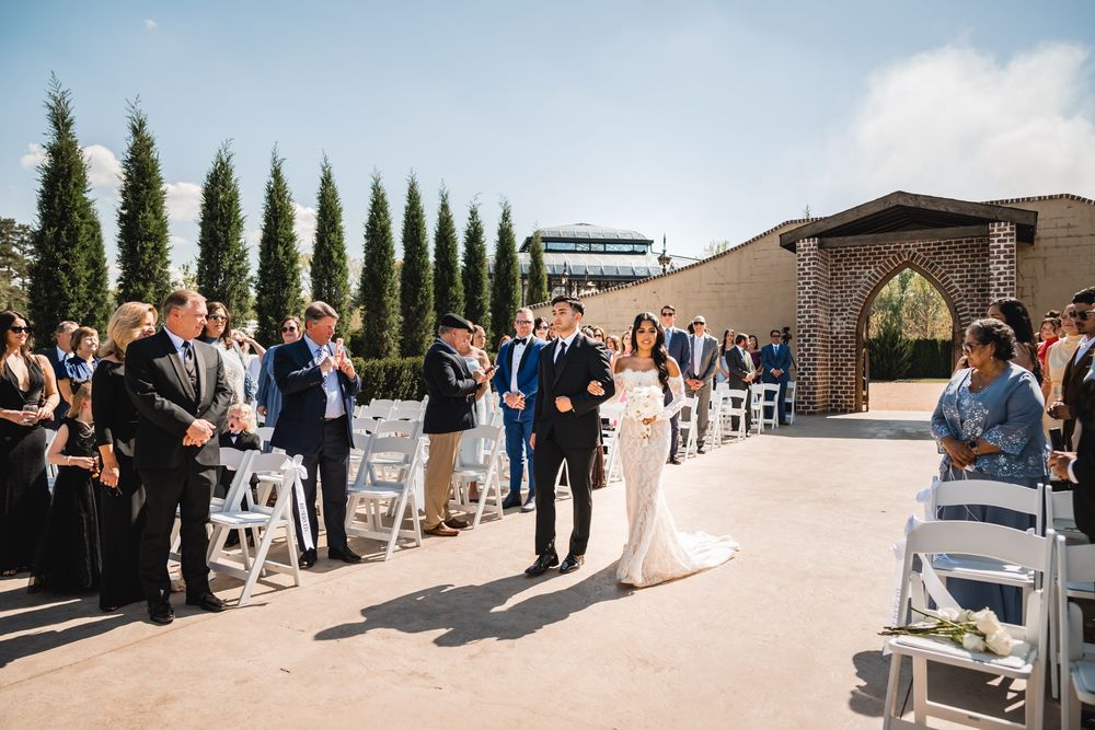 Bride walks down outdoor aisle with escort at elegant winery wedding ceremony on sunny day.