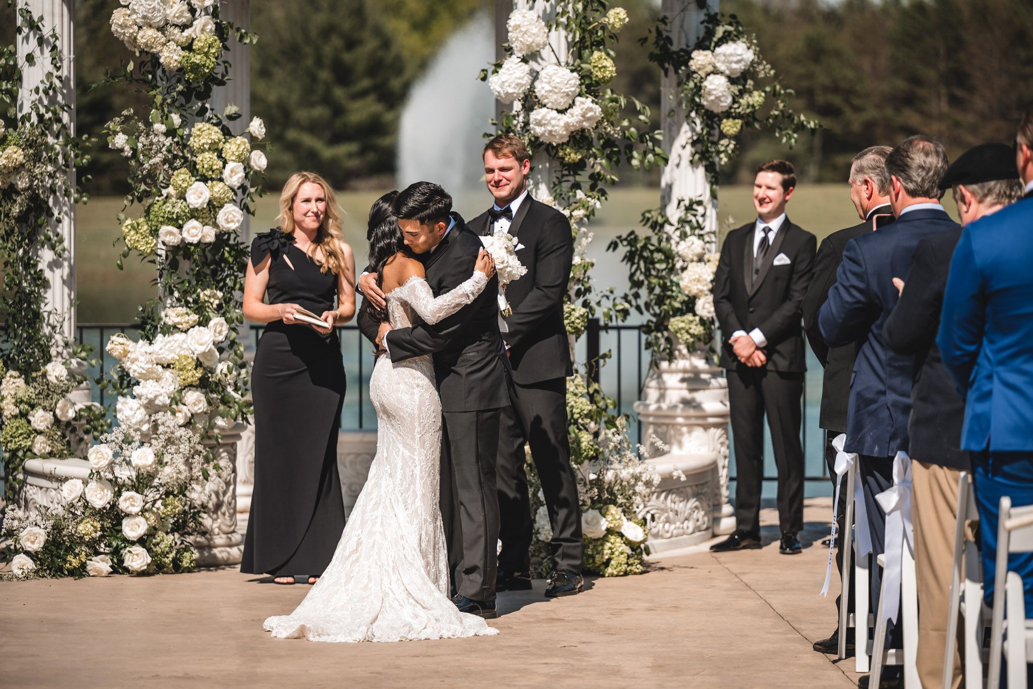Bride and groom share first kiss at floral arch altar with officiant and wedding party.