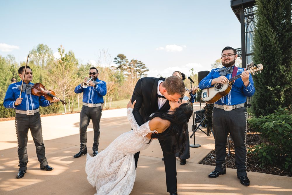 Groom dips bride for a kiss while a mariachi band in blue uniforms plays at an outdoor wedding celebration.
