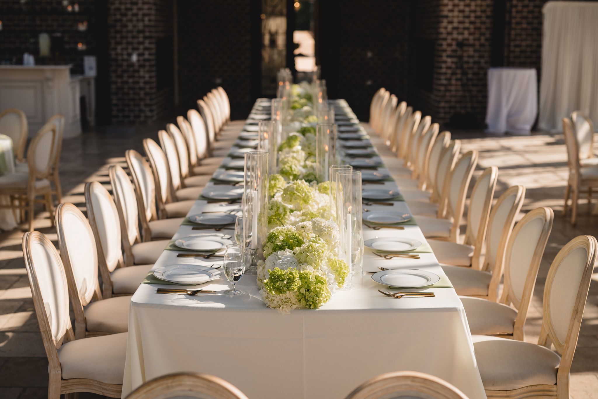 Long banquet table with white linens, green hydrangea centerpieces in brick-walled venue.