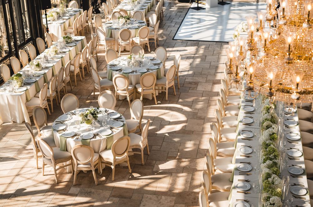 Elegant wedding reception with round tables, gold chairs, white linens, and floral centerpieces in a sunlit venue.