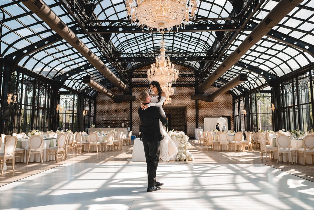 Groom lifts bride in stunning glass-ceiling ballroom with chandelier and reception tables.