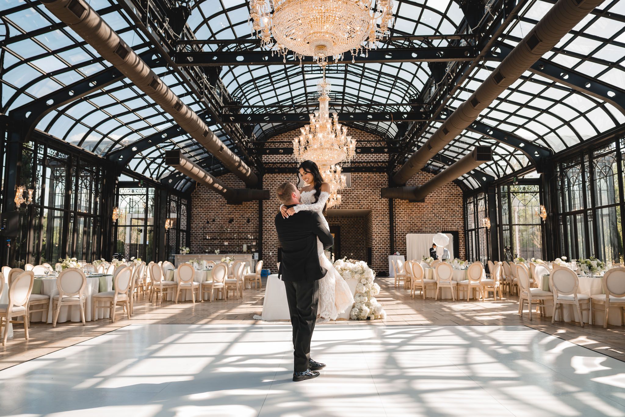 Groom lifts bride in stunning glass-ceiling ballroom with chandelier and reception tables.
