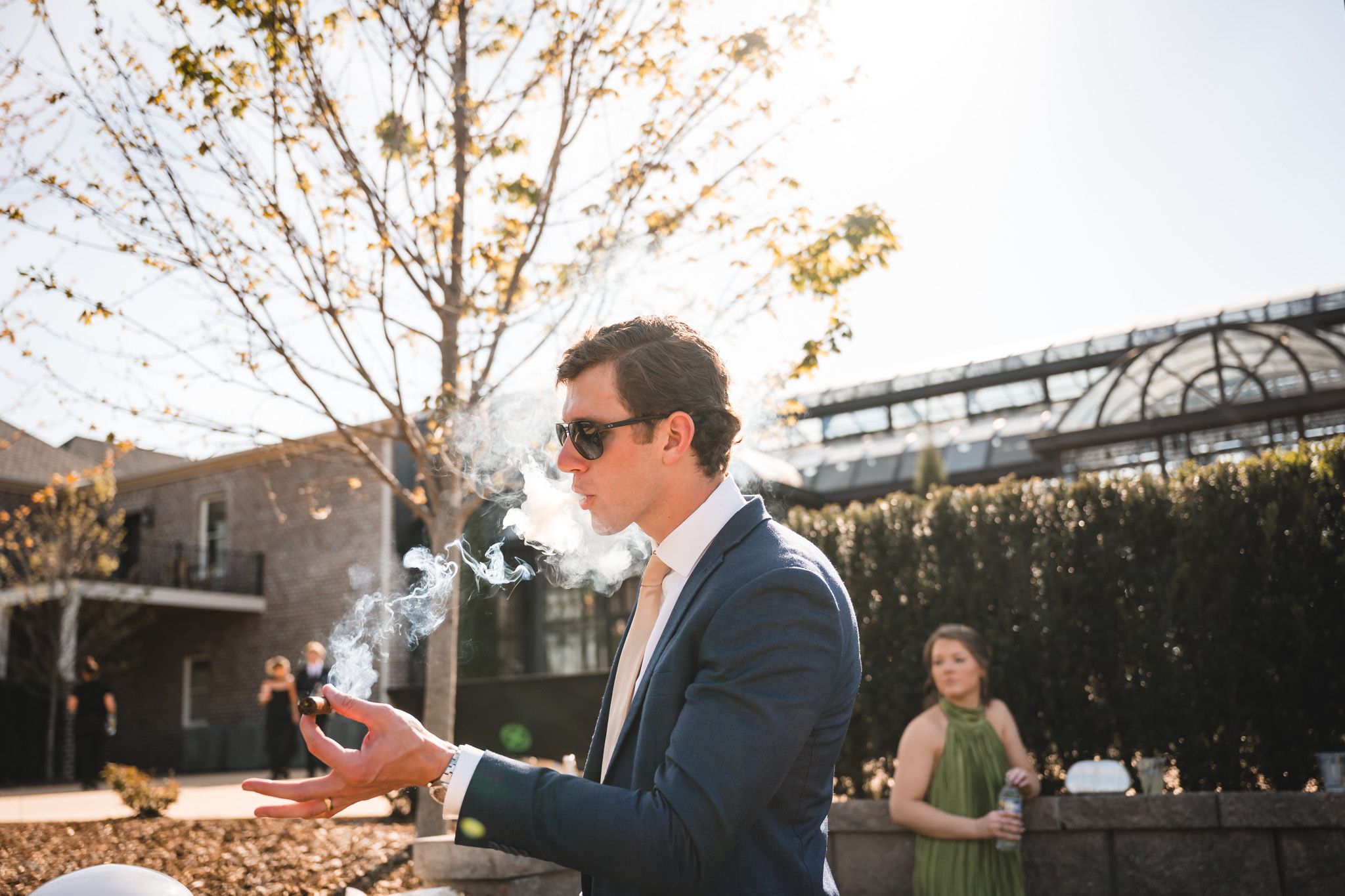 Wedding guest in blue suit pops champagne bottle outdoors at a sunny wedding reception.