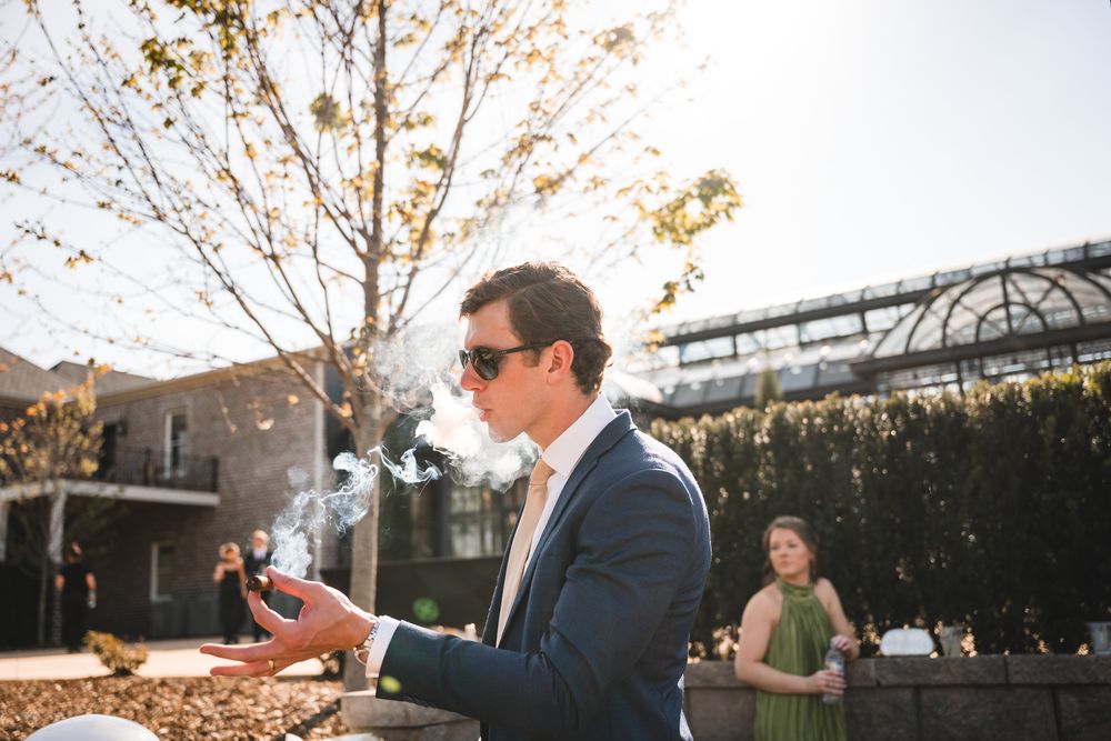 Wedding guest in blue suit pops champagne bottle outdoors at a sunny wedding reception.