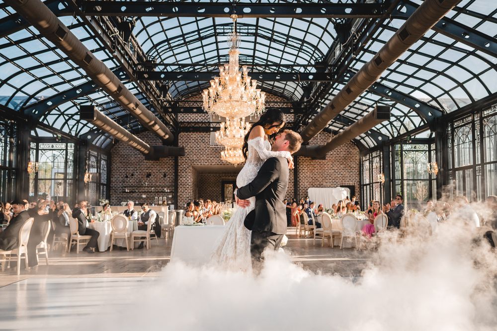 Couple shares first dance in stunning glass-ceiling ballroom with chandelier and dramatic fog effects.