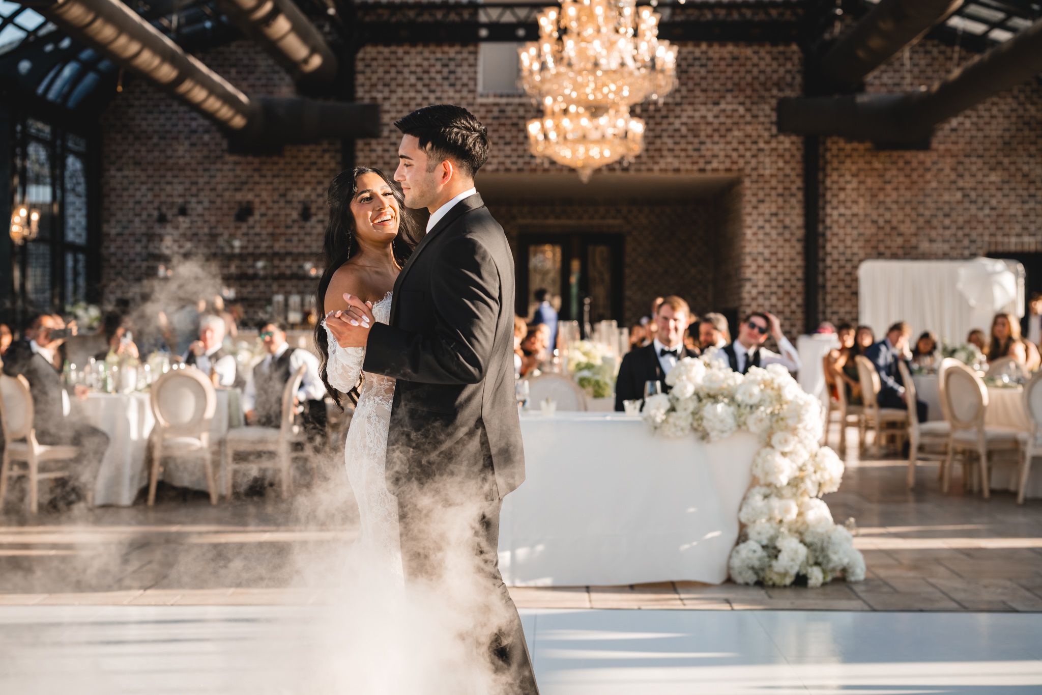 Bride and groom share first dance on foggy dance floor in elegant chandelier-lit wedding reception venue.