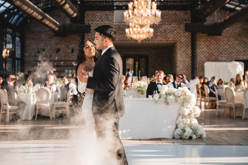 Bride and groom share first dance on foggy dance floor in elegant chandelier-lit wedding reception venue.