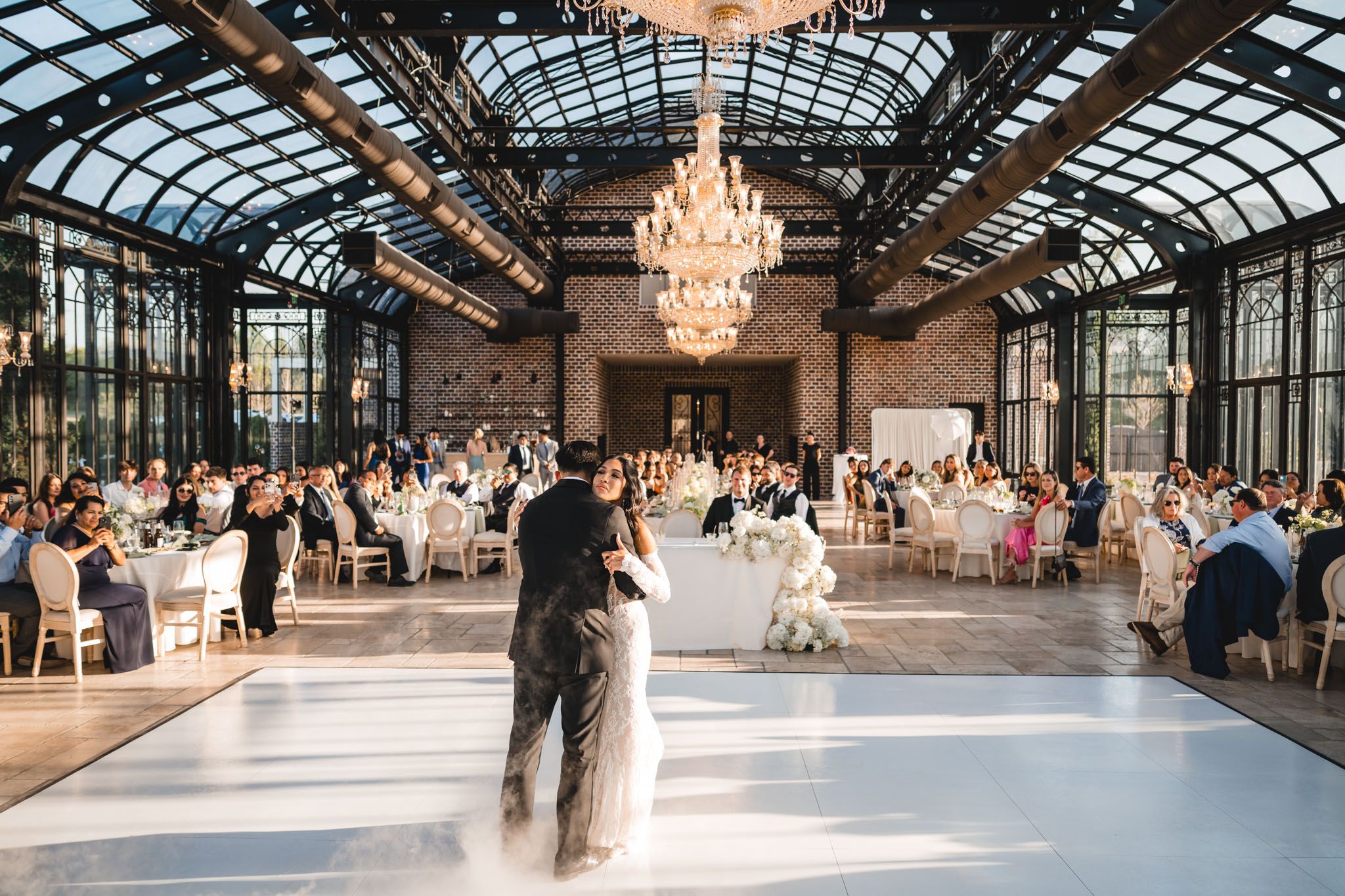 Couple shares first dance on white floor in elegant glass greenhouse wedding venue with chandeliers and seated guests.