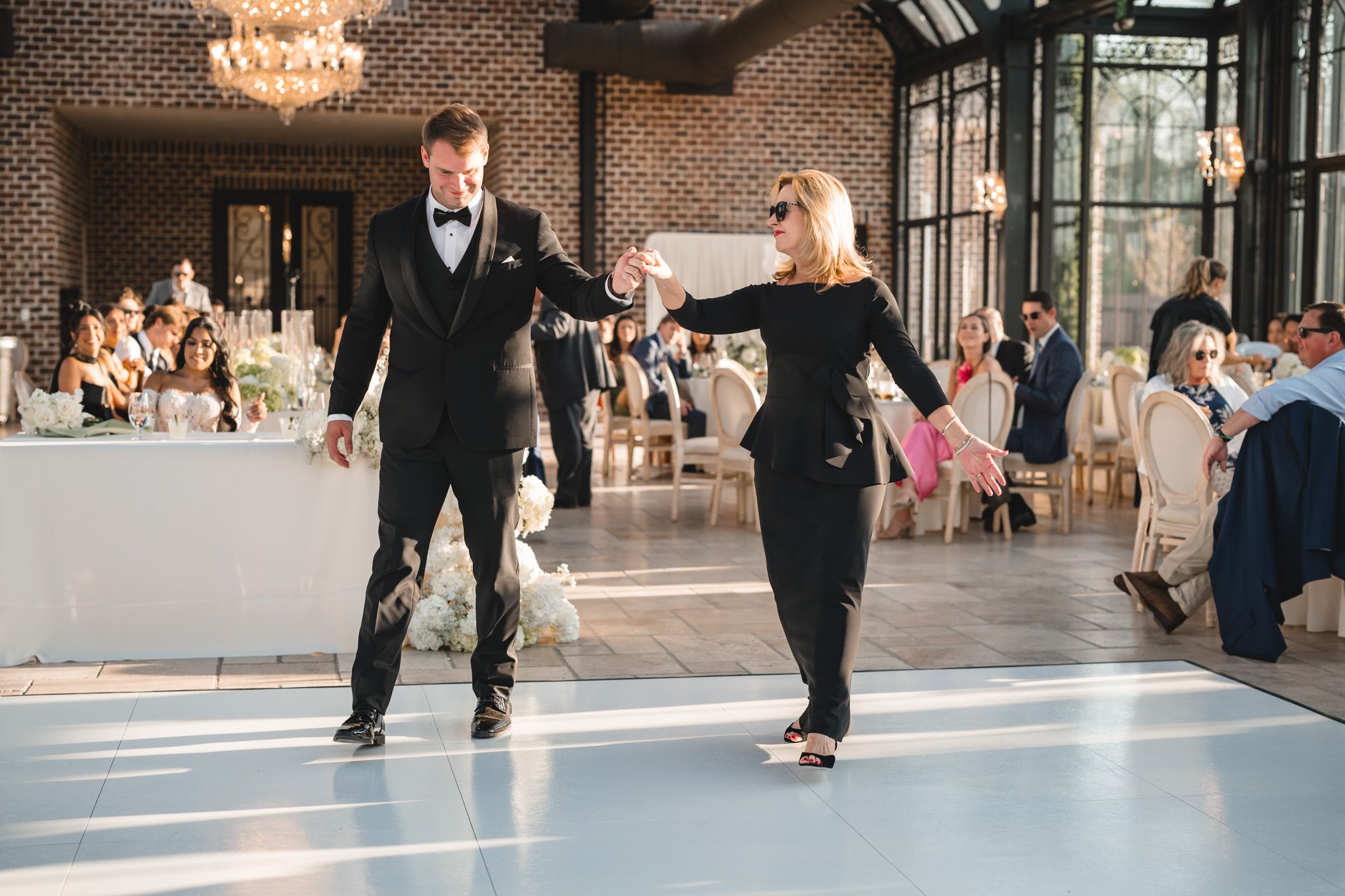 Groom dances with mother in black dress during wedding reception in elegant glass conservatory.