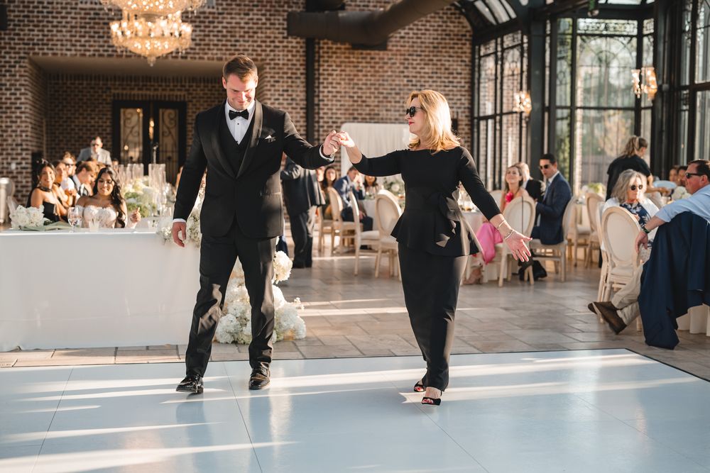 Groom dances with mother in black dress during wedding reception in elegant glass conservatory.