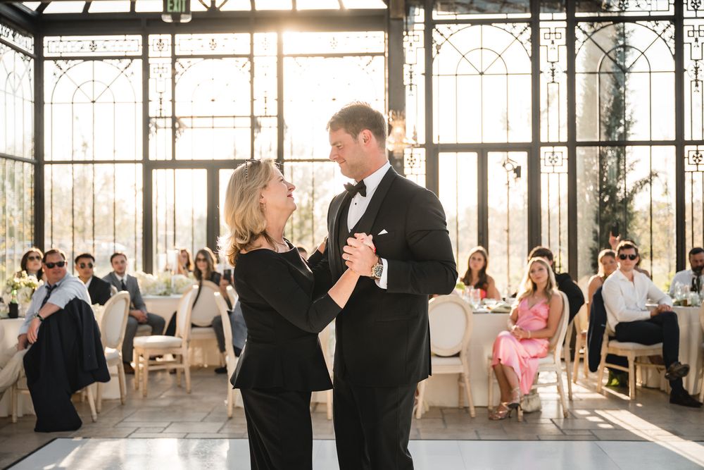 Mother and son share a dance in a sunlit greenhouse wedding venue with guests watching in the background.