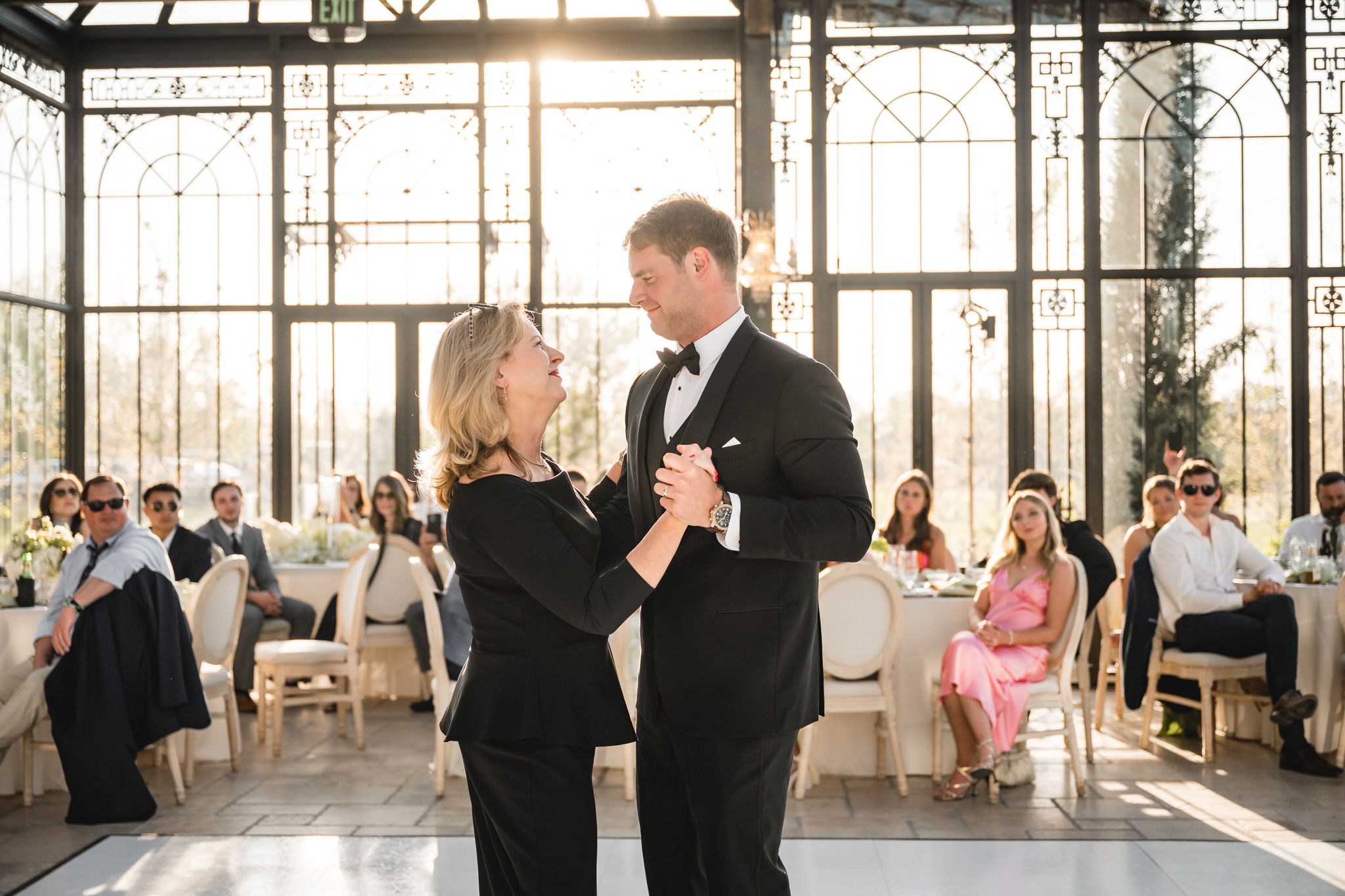 Mother and son share a dance in a sunlit greenhouse wedding venue with guests watching in the background.