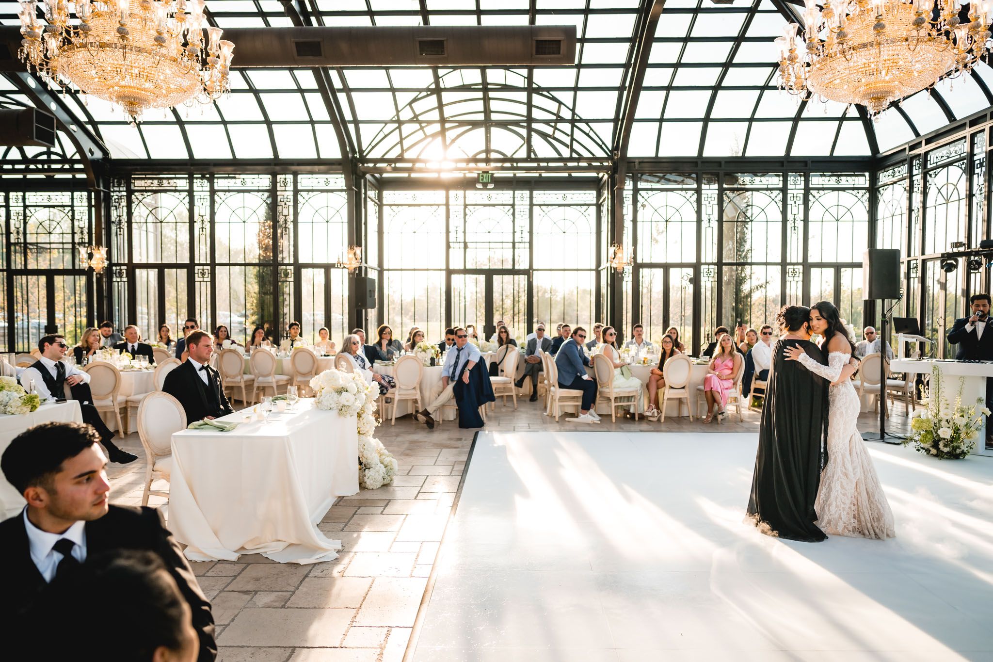 Elegant wedding reception in a glass greenhouse venue with guests seated at white-draped tables as couple shares first dance.